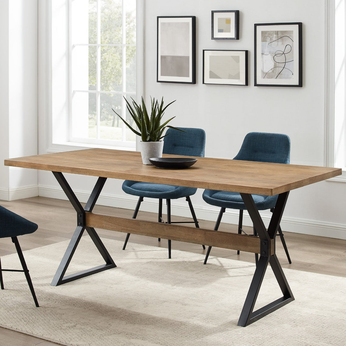 Wood and metal dining table with a rectangular wooden top and black metal X-shaped legs, paired with blue upholstered chairs on a light-colored rug, set in a bright room with abstract art and large windows.