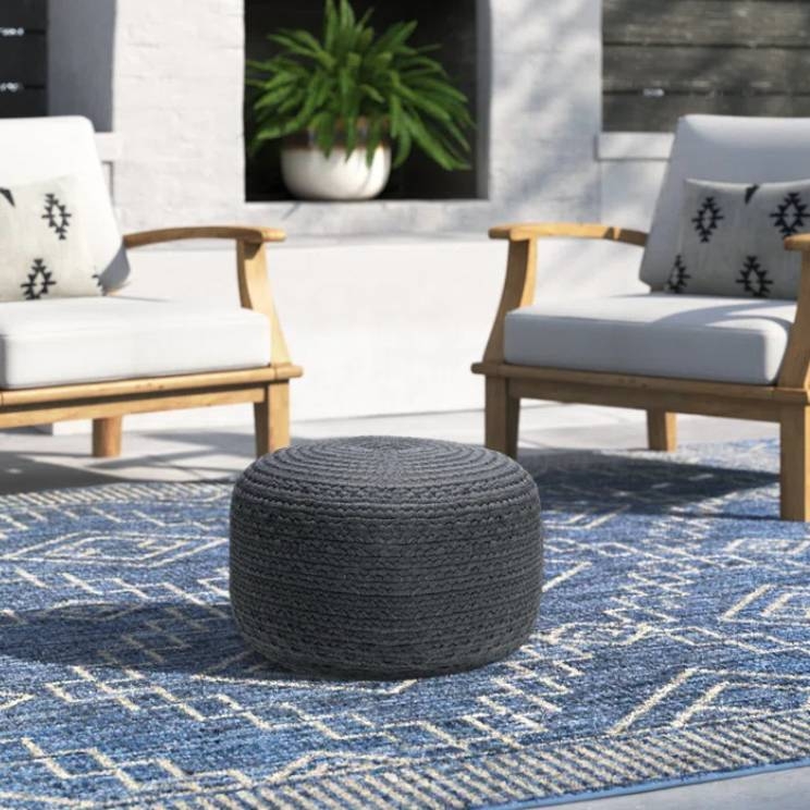 The Joss & Main Patio Ottoman is a round, woven pouf-style seat in a dark color, set on a patterned blue and white rug. It's accompanied by two wooden armchairs with white cushions and patterned pillows in a cozy outdoor setting.