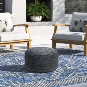 The Joss & Main Patio Ottoman is a round, woven pouf-style seat in a dark color, set on a patterned blue and white rug. It's accompanied by two wooden armchairs with white cushions and patterned pillows in a cozy outdoor setting.