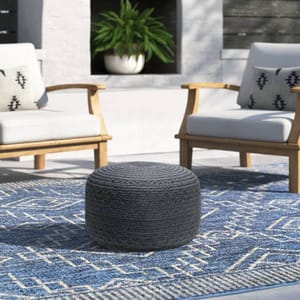 The Joss & Main Patio Ottoman is a round, woven pouf-style seat in a dark color, set on a patterned blue and white rug. It's accompanied by two wooden armchairs with white cushions and patterned pillows in a cozy outdoor setting.