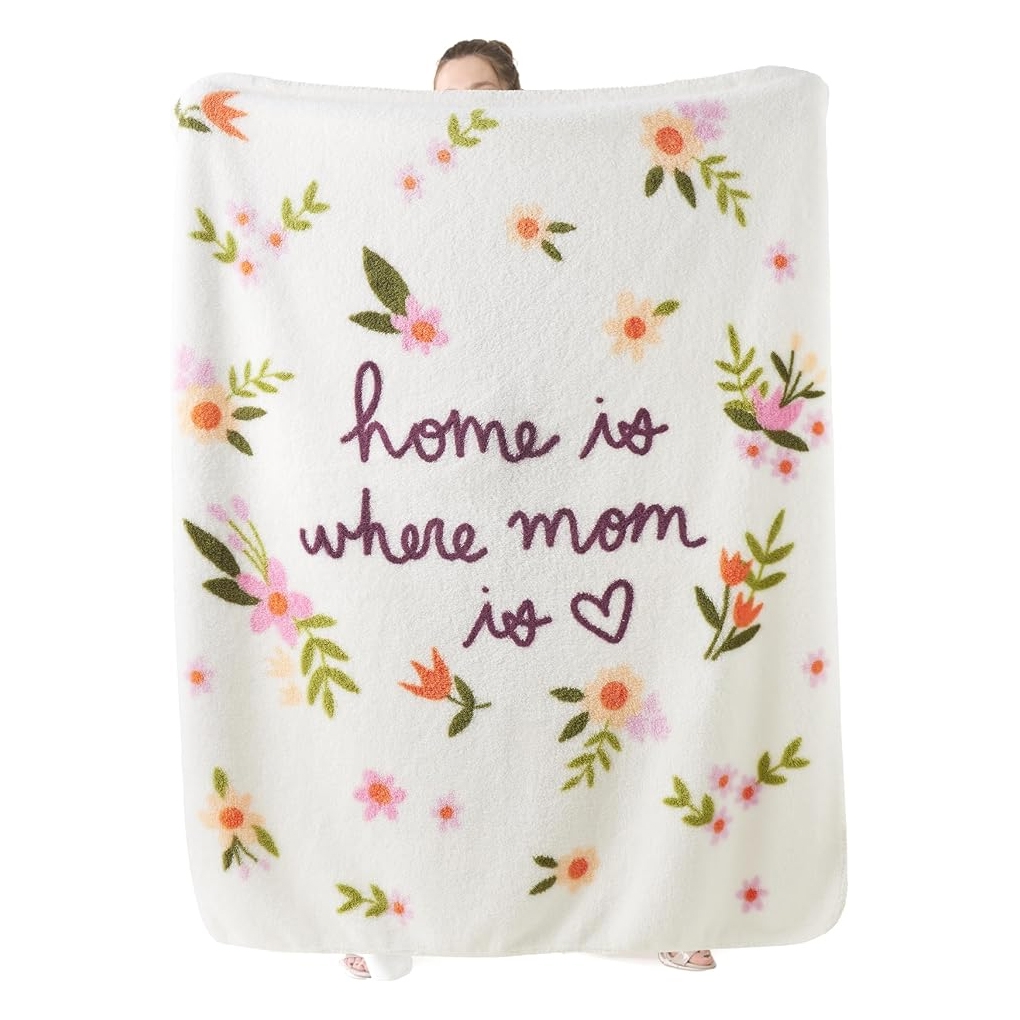 A white fleece blanket with floral designs features the phrase \“home is where mom is\“ in purple script, accompanied by a heart symbol.