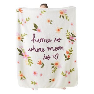A white fleece blanket with floral designs features the phrase \“home is where mom is\“ in purple script, accompanied by a heart symbol.