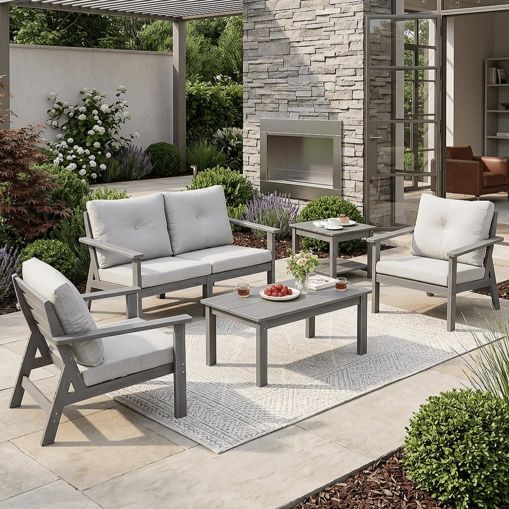 Outdoor patio set with a grey cushioned sofa, two chairs, and two tables on a rug, set in a garden with a stone wall and pergola.