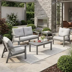 Outdoor patio set with a grey cushioned sofa, two chairs, and two tables on a rug, set in a garden with a stone wall and pergola.