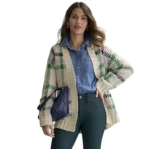 A woman models a beige and green plaid cardigan over a denim shirt, paired with dark green pants, holding a dark blue bag.