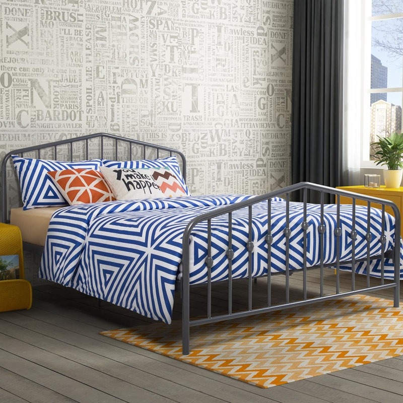 A bedroom features a metal bed frame with blue and white geometric bedding, complemented by decorative pillows with varied patterns. The backdrop includes a typographic wallpaper, a yellow nightstand, and a grey curtain, with a yellow and white chevron rug on the floor.