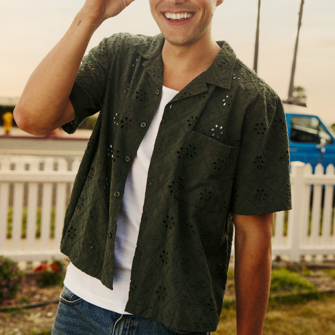 A person wears a short-sleeve, dark green shirt with eyelet patterns over a white T-shirt, paired with jeans.