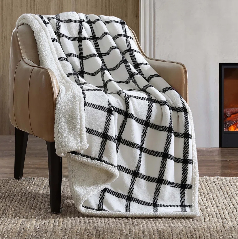 A cozy, black-and-white checkered sherpa fleece blanket is draped over a tan armchair. It is positioned on a woven rug next to a fireplace.