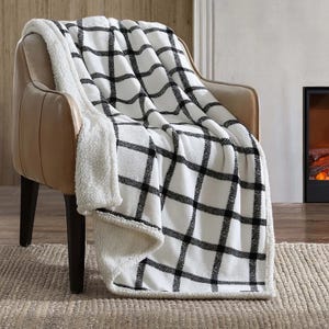 A cozy, black-and-white checkered sherpa fleece blanket is draped over a tan armchair. It is positioned on a woven rug next to a fireplace.