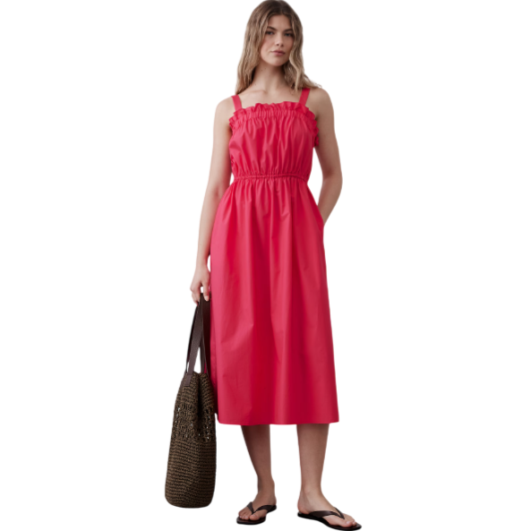 A woman is wearing a red sleeveless, ruffled midi dress with an elastic waist. She holds a woven brown tote bag and wears black flip-flops.