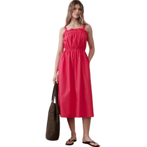 A woman is wearing a red sleeveless, ruffled midi dress with an elastic waist. She holds a woven brown tote bag and wears black flip-flops.