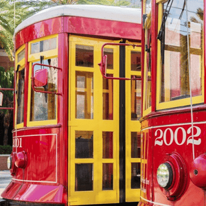 Two red and yellow streetcars are shown side by side, numbered 2001 and 2002, against a backdrop of palm trees.