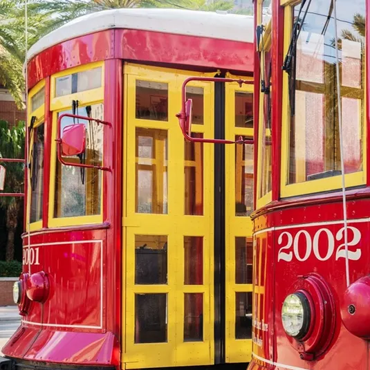 Two red and yellow streetcars are shown side by side, numbered 2001 and 2002, against a backdrop of palm trees.