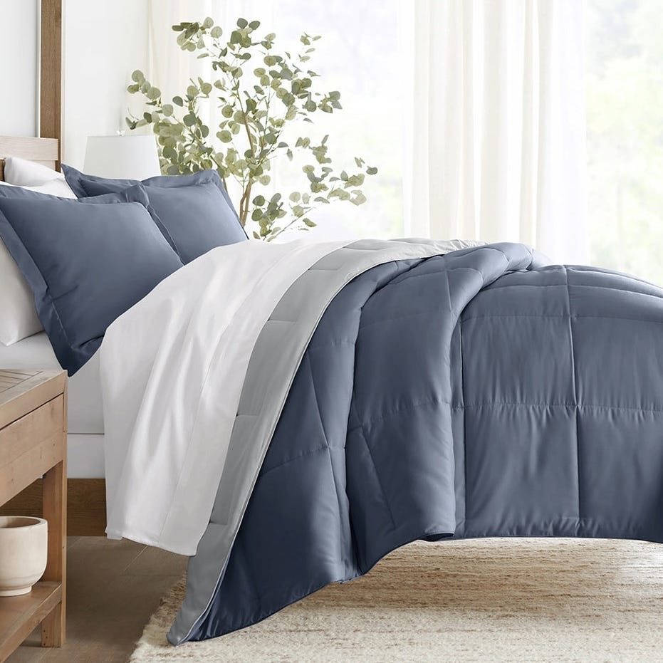 A cozy bedroom scene with a neatly made bed featuring a blue and grey comforter and matching pillows, accented by white sheets. A small bedside table holds a decorative item, and a plant adds a touch of greenery to the minimalist decor.