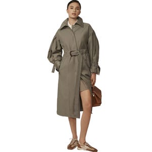 A person wears a belted trench coat with voluminous sleeves, paired with a short dress and brown-and-white sneakers, carrying a brown handbag.