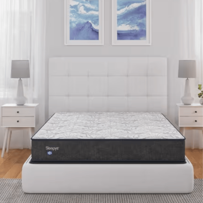 Sleepys mattress on a white upholstered bed frame with a quilted headboard, flanked by two white nightstands with lamps, set against a backdrop of two abstract blue wall art pieces.