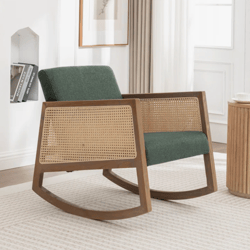 A wooden rocking chair with a green cushioned seat, featuring woven cane sides, is placed on a light-colored rug next to a wooden side table, within a room decorated with framed art and shelves displaying books.