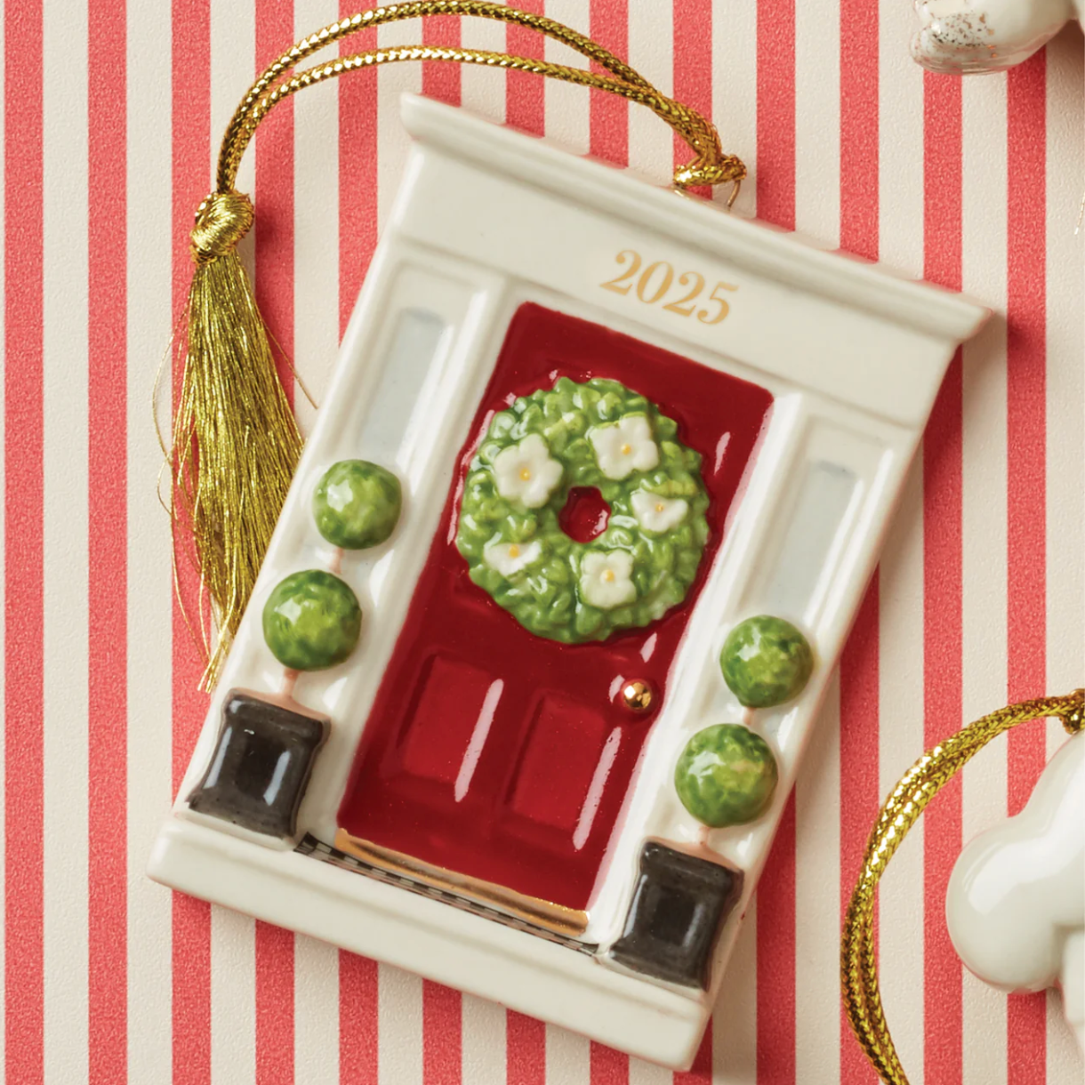 A ceramic ornament shaped like a red front door adorned with a wreath, labeled \“2025,\“ featuring decorative green topiaries and a gold tassel on a striped background.