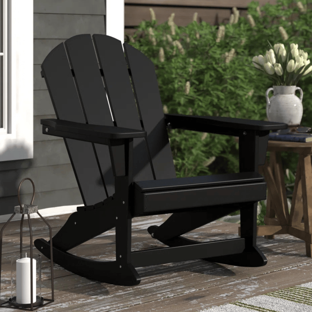 Black Adirondack rocking chair on a wooden deck, accompanied by a candle lantern and a vase of white flowers.