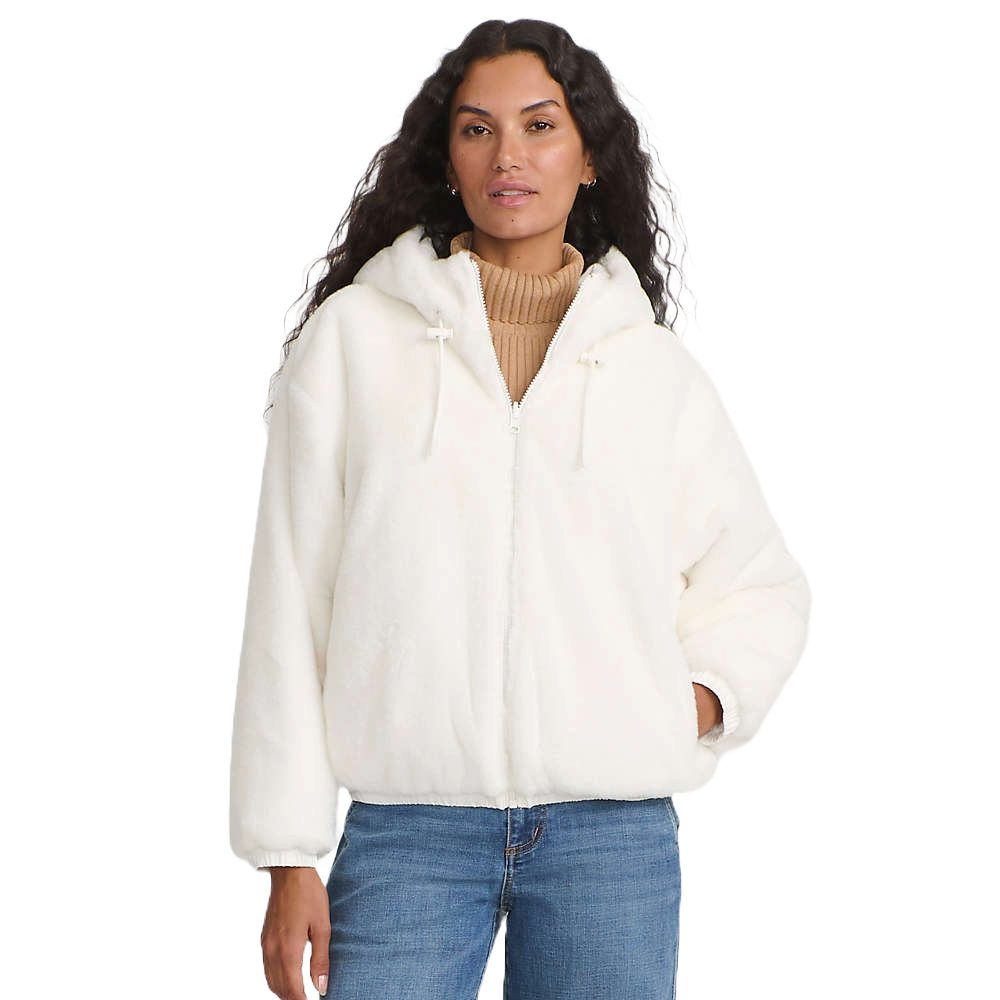 A woman wears a white, plush zip-up hoodie with a hood and drawstrings over a tan turtleneck sweater, paired with blue jeans.