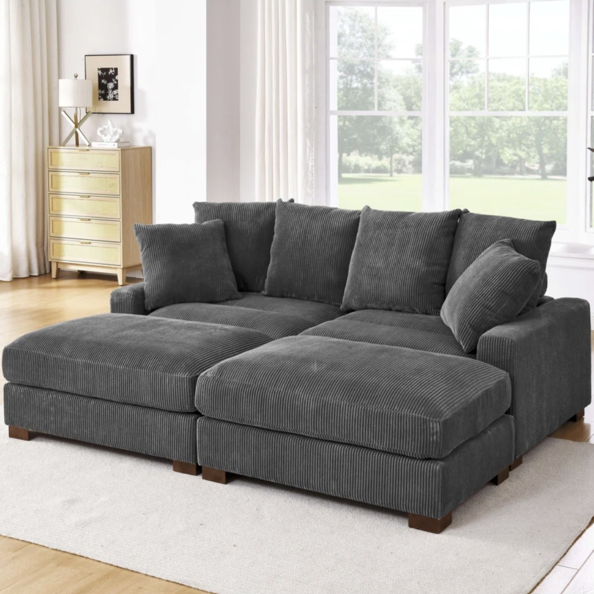 A gray corduroy sectional sofa with multiple cushions is placed on a light-colored rug in a bright, modern living room featuring large windows and wooden flooring. A light wood dresser and a potted plant are in the background.