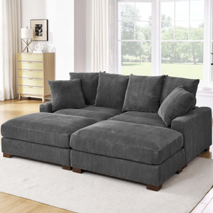 A gray corduroy sectional sofa with multiple cushions is placed on a light-colored rug in a bright, modern living room featuring large windows and wooden flooring. A light wood dresser and a potted plant are in the background.