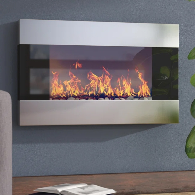 A sleek wall-mounted electric fireplace with a modern stainless steel finish featuring realistic flames and decorative pebbles. It's elegantly integrated into a living room setting with adjacent furniture and plants.