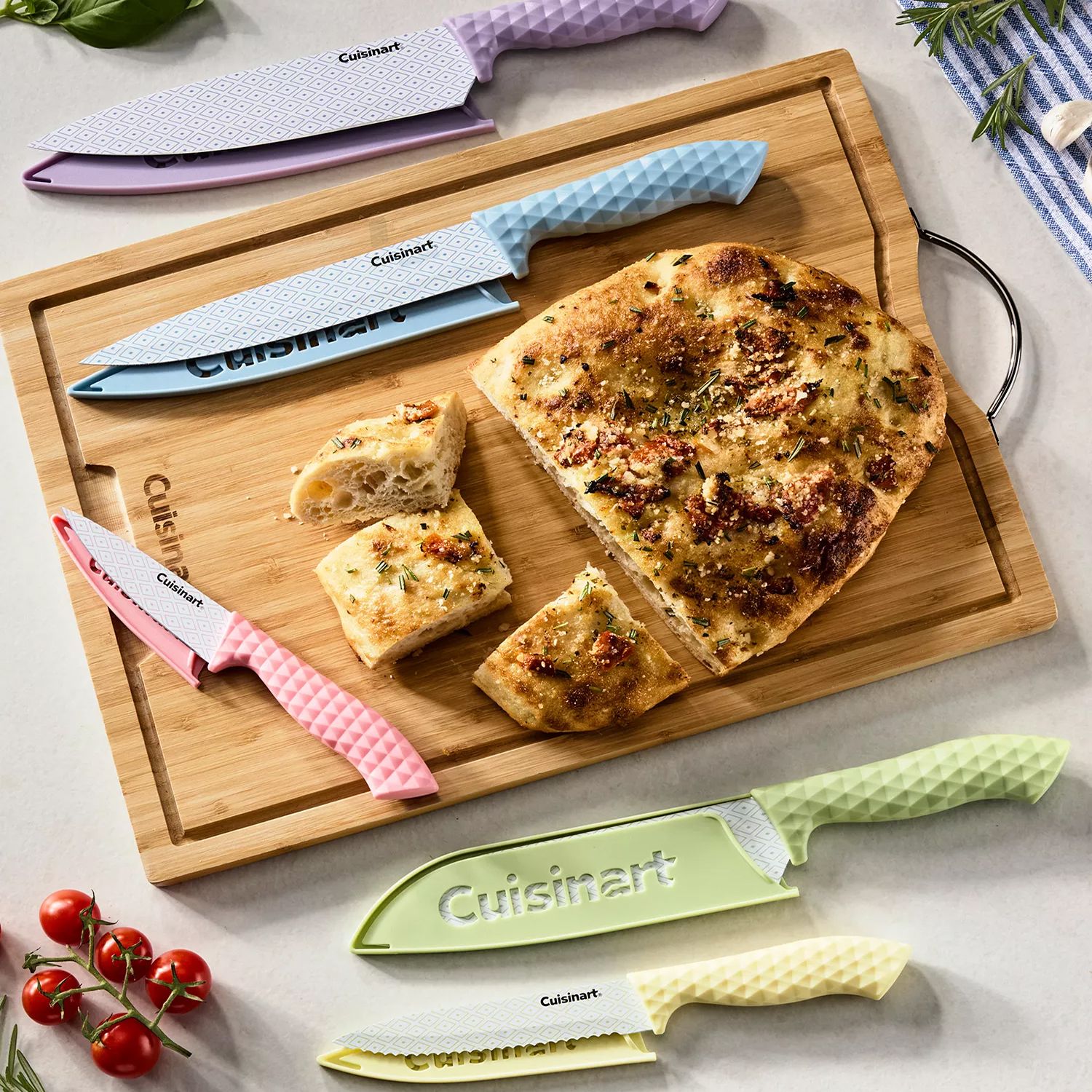 A wooden cutting board displays a sliced focaccia and a set of colorful Cuisinart knives with textured handles in pink, blue, yellow, green, and purple. Cherry tomatoes and rosemary are also visible.