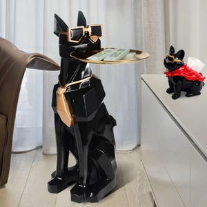 A geometric black dog sculpture with sunglasses and a gold tie holds a tray with money on it, while a smaller dog sculpture wearing a red cape and sunglasses sits on a cabinet.