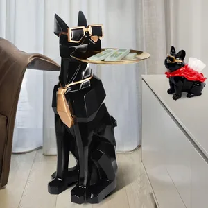A geometric black dog sculpture with sunglasses and a gold tie holds a tray with money on it, while a smaller dog sculpture wearing a red cape and sunglasses sits on a cabinet.