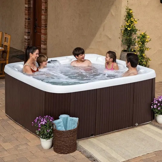 An outdoor hot tub with a brown exterior and white interior, featuring people enjoying the warm water. The setup includes potted plants, a basket of rolled towels, and a decorative rug on the patio.