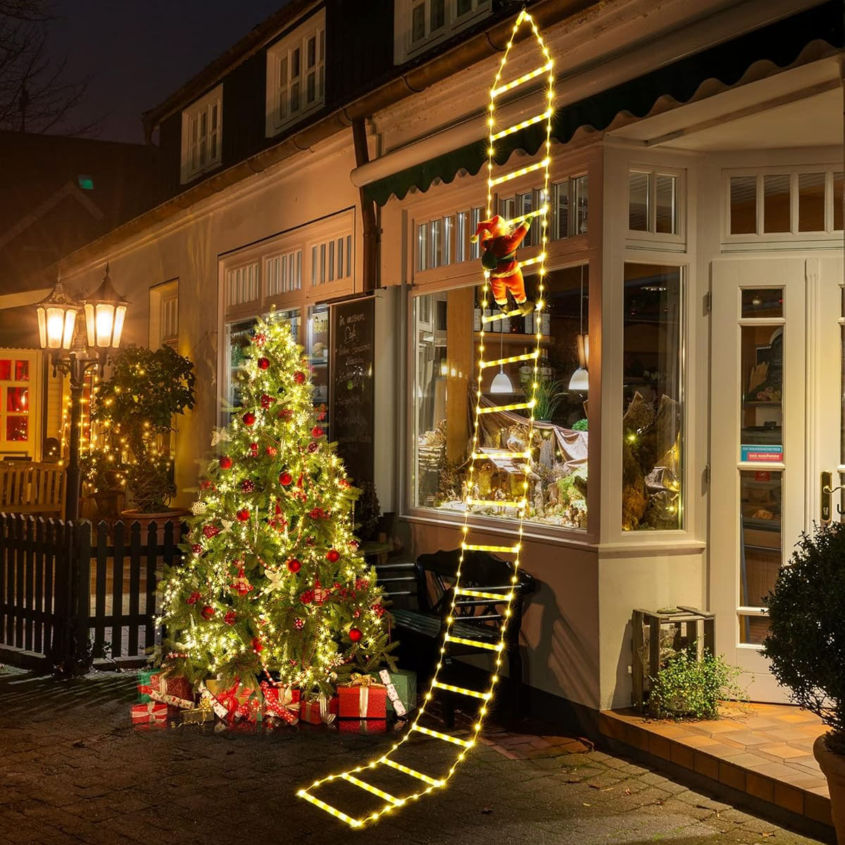 A 10-foot LED ladder with warm white lights is attached to a building, featuring a Santa figure climbing up. Nearby, a decorated Christmas tree with lights and presents enhances the festive exterior scene.