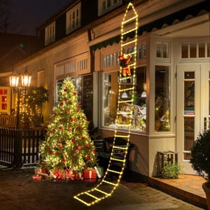 A 10-foot LED ladder with warm white lights is attached to a building, featuring a Santa figure climbing up. Nearby, a decorated Christmas tree with lights and presents enhances the festive exterior scene.