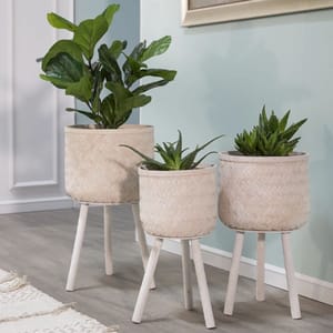 Three round woven bamboo and pine wood planters in varying heights, each with three white legs. The planters contain different green plants, adding a stylish and natural touch to the decor.
