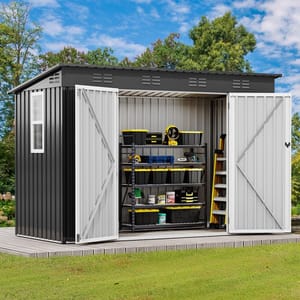 A metal storage shed with open double doors displays shelves stacked with toolboxes, paint cans, and a ladder, set on a wooden platform in a grassy area surrounded by trees.
