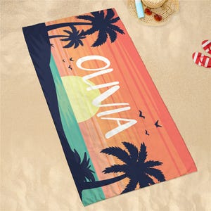 Personalized quick-dry beach towel with \“Olivia\“ in white text on a sunset background featuring silhouettes of palm trees and birds. It's displayed on sand with a hat, sunscreen, sunglasses, and flip-flops nearby.
