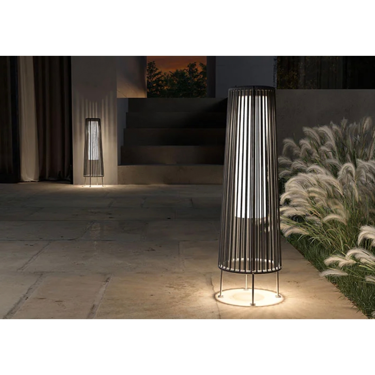 Two vertical outdoor floor lamps with cylindrical designs and metal frames, illuminating a patio area at dusk.