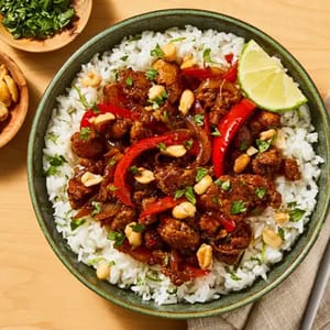 A bowl of white rice topped with stir-fried meat, red bell peppers, peanuts, and garnished with fresh herbs and a lime wedge.
