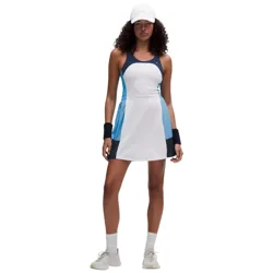 A woman models a sleeveless, color-blocked tennis dress with a white base, blue and black accents, paired with white sneakers, wristbands, and a white cap.