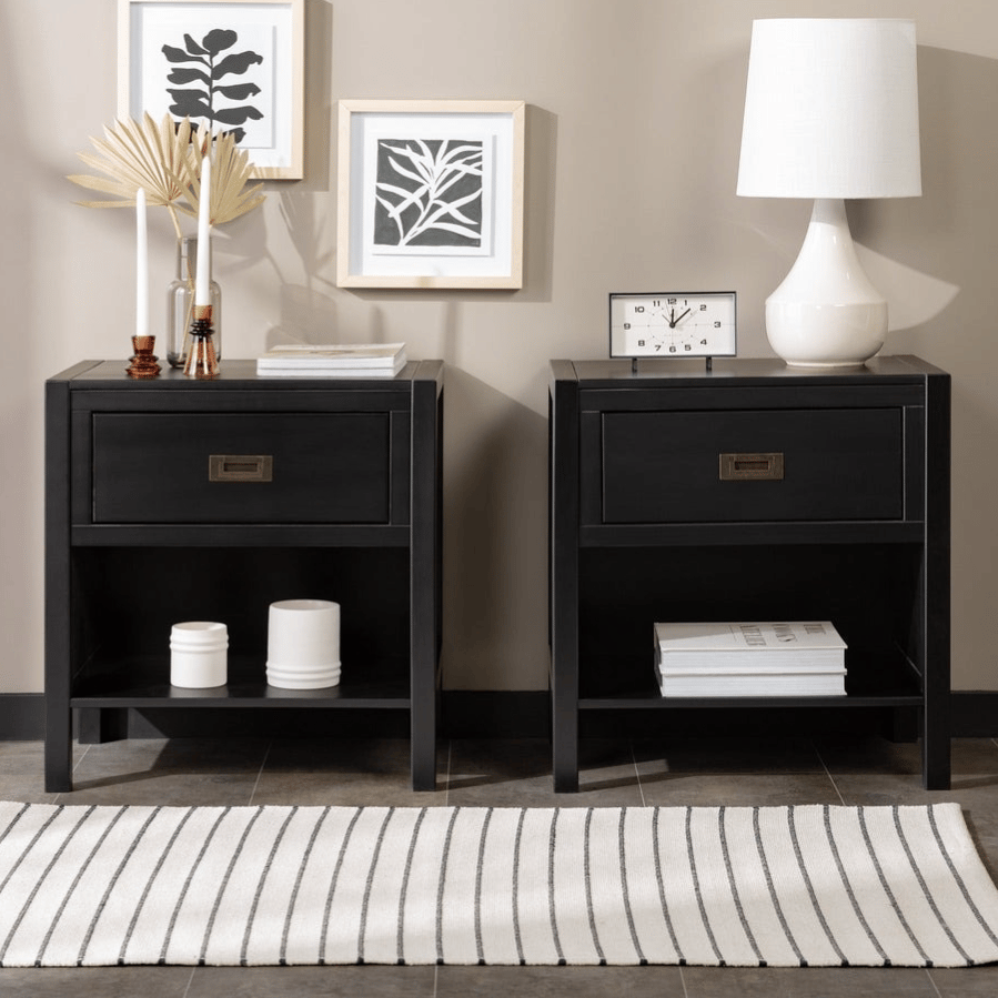 Two black nightstands with drawers; one adorned with a clock and lamp, the other with candles and decor. Wall features minimalist leaf art, and there's a striped rug on the floor.