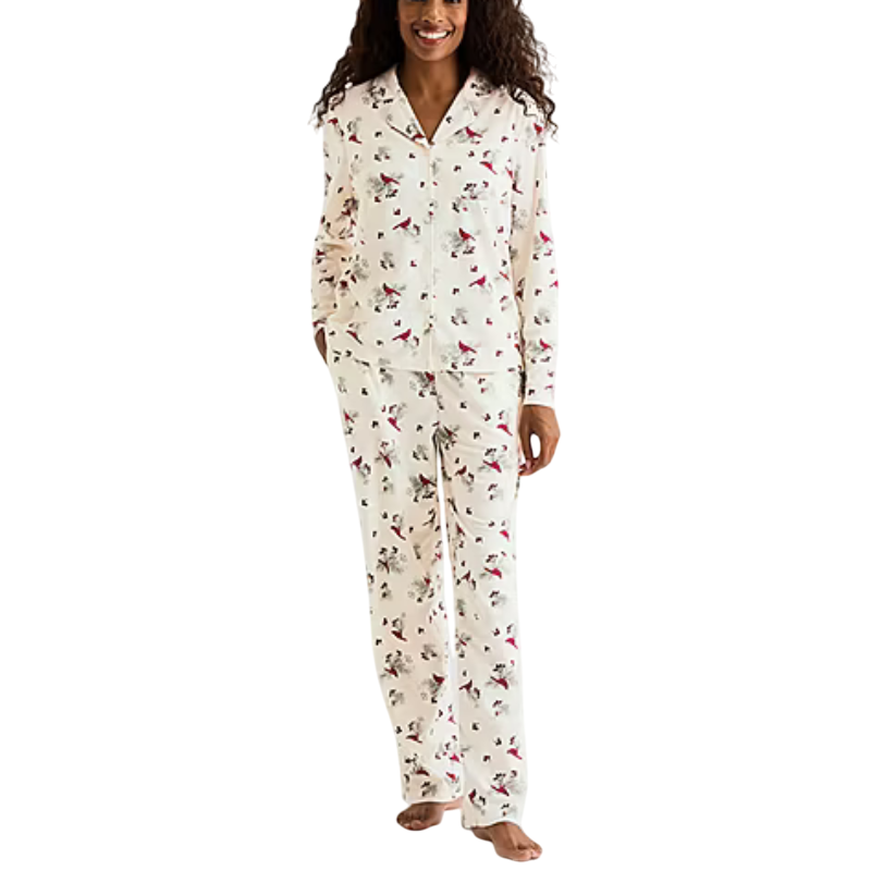 A woman is wearing a cream-colored pajama set with a floral and bird print, featuring a long-sleeve button-up top and matching pants.