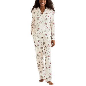 A woman is wearing a cream-colored pajama set with a floral and bird print, featuring a long-sleeve button-up top and matching pants.