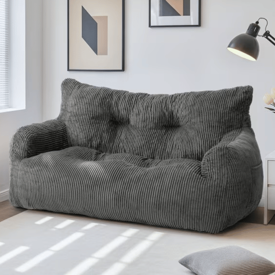 A dark grey corduroy sofa is placed in a well-lit room, accompanied by minimalistic wall art, a floor lamp, and a small side table with flowers. A matching cushion is on the floor.