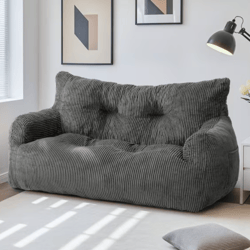 A dark grey corduroy sofa is placed in a well-lit room, accompanied by minimalistic wall art, a floor lamp, and a small side table with flowers. A matching cushion is on the floor.