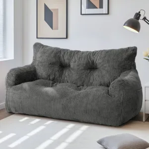 A dark grey corduroy sofa is placed in a well-lit room, accompanied by minimalistic wall art, a floor lamp, and a small side table with flowers. A matching cushion is on the floor.