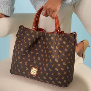 A brown Dooney & Bourke tote bag with a monogram pattern and brown leather handles, held by a person wearing a light blue sweater and white pants, standing next to a plush, cream-colored chair.