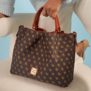A brown Dooney & Bourke tote bag with a monogram pattern and brown leather handles, held by a person wearing a light blue sweater and white pants, standing next to a plush, cream-colored chair.