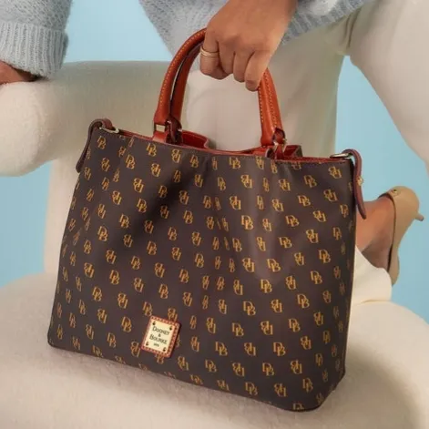 A brown Dooney & Bourke tote bag with a monogram pattern and brown leather handles, held by a person wearing a light blue sweater and white pants, standing next to a plush, cream-colored chair.