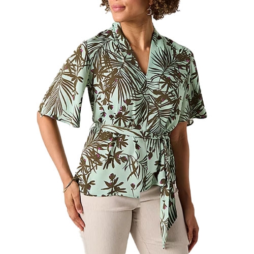 A woman wearing a short-sleeve, green and brown floral wrap blouse with a tie at the waist, paired with light-colored pants.