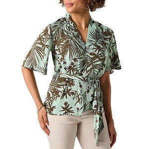 A woman wearing a short-sleeve, green and brown floral wrap blouse with a tie at the waist, paired with light-colored pants.